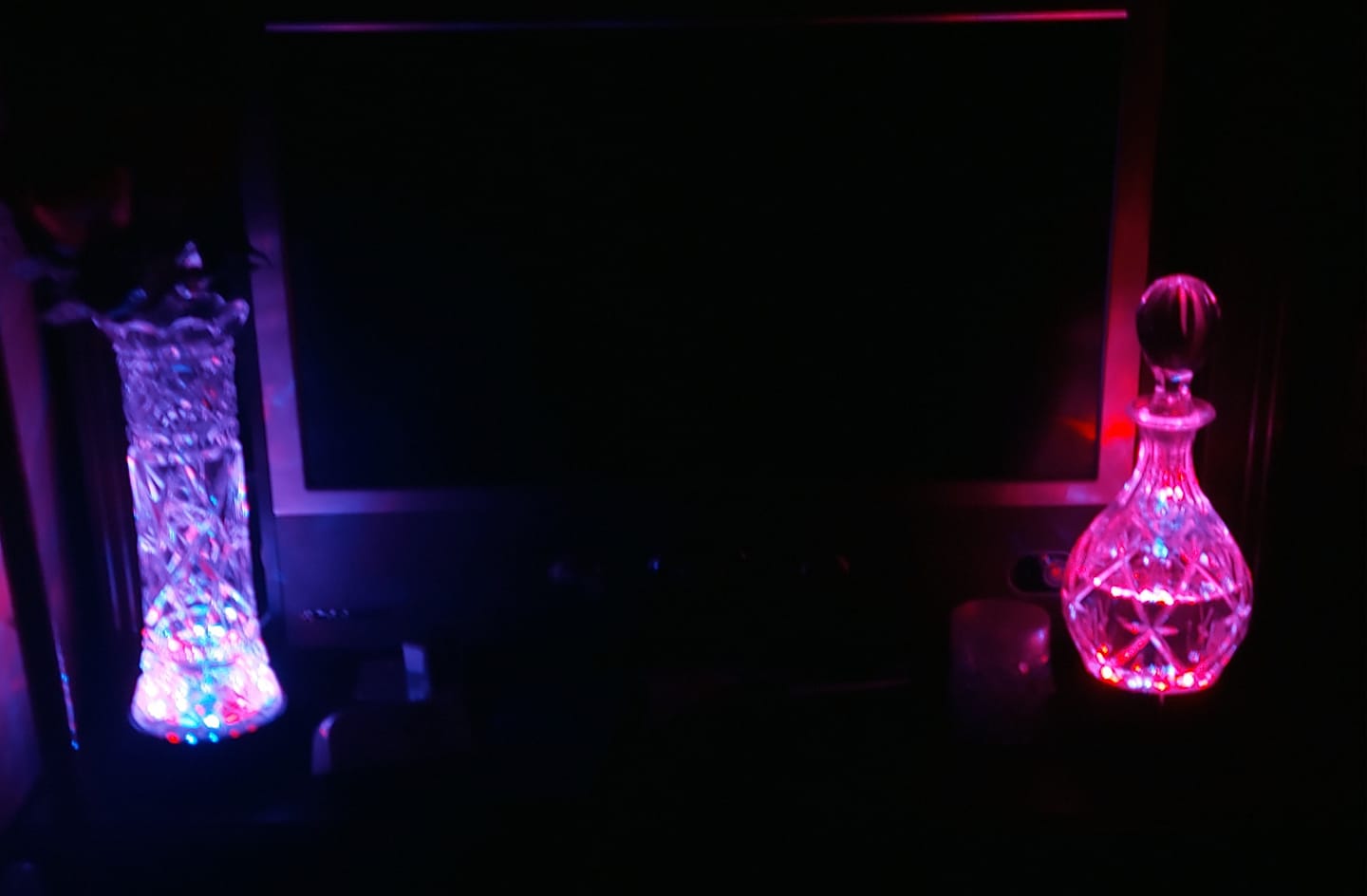 A photo taken in the dark of a decanter illuminated by magenta light and a vase illuminated by purple